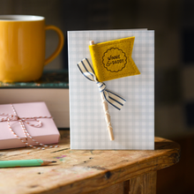 Personalised Father’s Day card with mustard felt flag reading “Henry & Daddy”, handmade keepsake card with ribbon and wooden stick detail. The card stands on a wooden table beside a book, mug, candle, and wrapped gift.