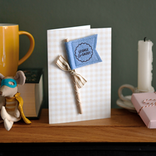 Personalised Father’s Day card with soft blue felt flag reading “George & Daddy”, handmade keepsake card with ribbon and wooden stick detail. The card stands on a wooden mantle beside a book, mug, candle, and wrapped gift.