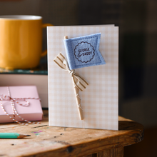 Personalised Father’s Day card with soft blue felt flag reading “George & Daddy”, handmade keepsake card with ribbon and wooden stick detail. The card stands on a wooden table beside a book, mug, candle, and wrapped gift.
