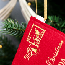 A red felt personalised Christmas envelope with a golden sleigh stamp and a Christmas tree postmark is hanging on a decorated Christmas tree with golden ribbon.