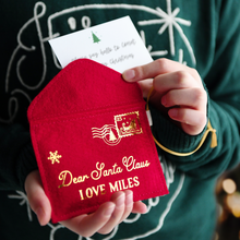 A child wearing a deep green jumper is holding a handmade red felt Christmas envelope that reads "Dear Santa Claus, Love Miles," with a festive letter card inside.