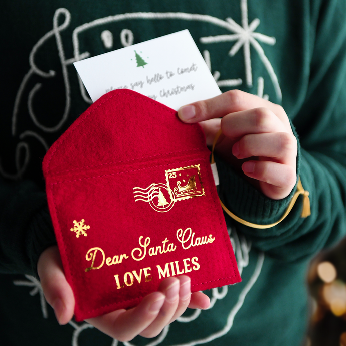 A child wearing a deep green jumper is holding a handmade red felt Christmas envelope that reads 