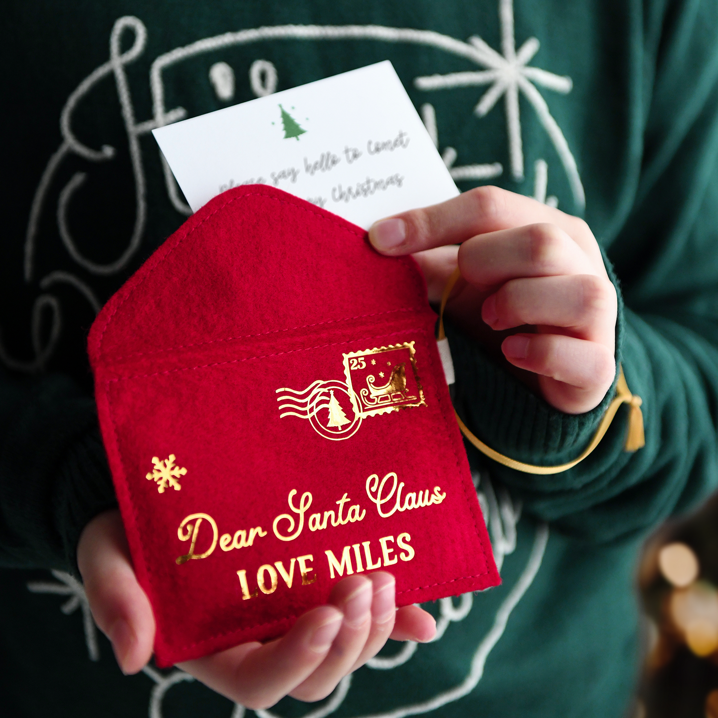 A child wearing a deep green jumper is holding a handmade red felt Christmas envelope that reads 