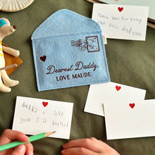 A handmade soft blue felt envelope with brown wording that reads 'Dearest Daddy, love Maude' lays on a dark green fabric surface. A young child is writing a notecard to Daddy beside the envelope. Several other mini notecards are around the envelope too.