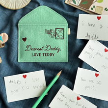 A handmade green felt envelope with the personalisation 'Dearest Daddy, love Maude' and a paper aeroplane stamp with postmark, lays on dark blue fabric. Notes to Daddy are written on the included heart notecards.