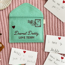 A handmade green felt envelope with the personalisation 'Dearest Daddy, love Maude' and a paper aeroplane stamp with postmark, lays on pink fabric. Notes to Daddy are written on the included heart notecards.
