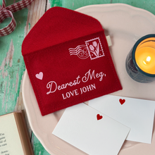 A handmade personalised red felt love letters envelope with the words 'Dearest Meg, love John' is displayed on a vintage-style plate with small heart cards, styled on a rustic green wooden table for Valentine’s Day.
