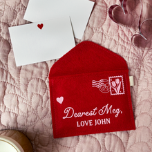 A handmade red felt envelope with a pale pink stamp and postmark design, and the words 'Dearest Meg, love John' rests on a pink quilt. Two notecards with small hearts on are tucked under the top of the envelope.