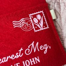 A handmade red felt envelope lays on a pale pink quilt. A pale pink stamp and postmark is in the top right corner, the stamp features a tulip design and the postmark is a bow design.