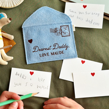 A handmade soft blue felt envelope with brown wording that reads 'Dearest Daddy, love Maude' lays on a dark green fabric surface. A young child is writing a notecard to Daddy beside the envelope. Several other mini notecards are around the envelope too.