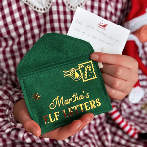 A child wearing a red gingham dress is holding a handmade green felt envelope with golden words reading "Martha's elf letters" and a candy cane design stamp in the corner. 