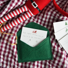 A handmade green Christmas envelope with a festive card tucked inside, written with a note to the elves, lays on a gingham cloth next to a Christmas elf toy.