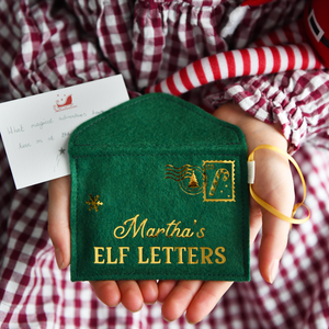 A child wearing a red gingham dress is holding a handmade green felt envelope with golden words reading "Martha's elf letters" and a candy cane design stamp in the corner. 