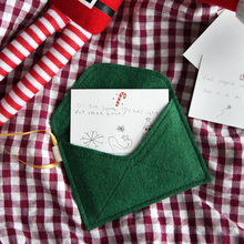 A handmade green Christmas envelope with a festive card tucked inside, written with a note to the elves, lays on a gingham cloth next to a Christmas elf toy.