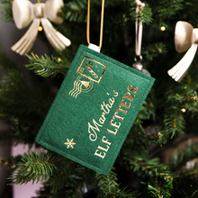A green felt personalised envelope decoration with golden words reading "Martha's Elf Letters" and a candy cane design stamp in the corner, hangs on a decorated Christmas tree.