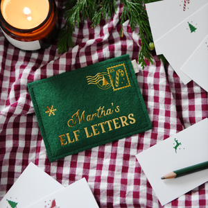 Green felt personalised envelope reading "Martha’s Elf Letters," placed on a red gingham cloth with festive notecards, a green pencil, greenery, and a lit candle.