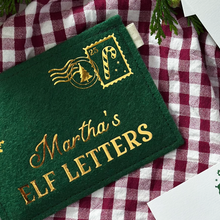 A green felt reusable envelope with the words "Martha's Elf Letters" in golden writing and a candy cane stamp in the top right corner, lays on red gingham fabric.