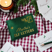 Green felt personalised envelope reading "Martha’s Elf Letters," placed on a red gingham cloth with festive notecards each featuring letters to the elves, with a green pencil, greenery, and a lit candle surrounding them.