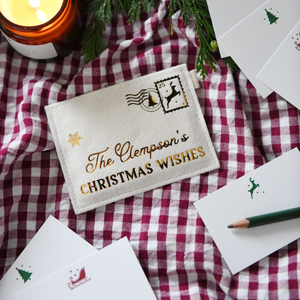 A cream felt personalised envelope reading "The Clempson's Christmas Wishes," placed on a red gingham cloth with festive notecards, a green pencil, greenery, and a lit candle.