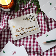 A cream felt personalised envelope reading "The Clempson's Christmas Wishes," placed on a red gingham cloth with festive notecards, a green pencil, greenery, and a lit candle.