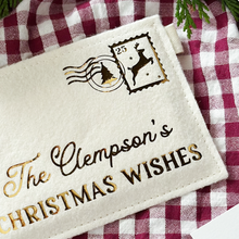 A cream felt personalised envelope reading "The Clempson's Christmas Wishes," and featuring a golden reindeer postage stamp is placed on a red gingham cloth.