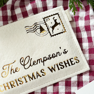A cream felt personalised envelope reading "The Clempson's Christmas Wishes," and featuring a golden reindeer postage stamp is placed on a red gingham cloth.
