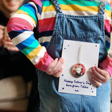 A young child wearing a striped top and blue dungarees holds a personalised Father's Day card behind her back. The card has a wooden photo decoration attached and the words Happy Father's Day love from Tilda xx