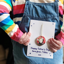 A young child wearing a striped top and blue dungarees holds a personalised Father's Day card behind her back. The card has a wooden photo decoration attached and the words Happy Father's Day love from Tilda xx