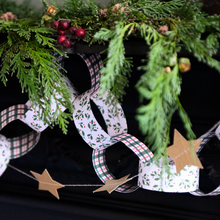 A close-up of a holiday garland with white paper rings printed with green holly leaves, red-and-green plaid edges, and small tan paper stars, styled with evergreen branches and red berries against a dark background.