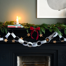 A festive Christmas garland draped over a black fireplace mantel, featuring white paper chain links with blue gingham, black-and-white checkered, and solid white patterns, accented with gold star cutouts and a red ribbon bow. The mantel is decorated with green pine garland, a lit white candle, and books against a dark gray wall.
