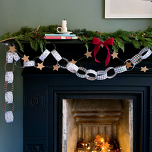 A decorative Christmas paper chain garland with patterned paper links and wooden stars hangs across a dark fireplace mantel, styled with evergreen branches, a red bow, books, and a candle in a cosy indoor setting.