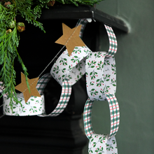 A Christmas-themed paper chain garland with white loops printed with green holly leaves and red berries, alternating with white loops featuring a red and green plaid pattern, and two brown paper star cutouts. The garland is hanging in front of a dark fireplace mantel decorated with evergreen branches.