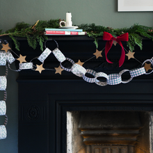 A festive Christmas paper chain garland with star-shaped links draped across a dark fireplace mantel, decorated with green pine garland, a red ribbon bow, and small books and a candle on top.