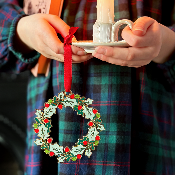 A child wearing a tartan dress holds a vintage-style enamel Christmas wreath ornament with green holly leaves and red berries, hanging from a red ribbon, alongside a small white candleholder with a lit candle.