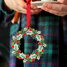 A child wearing a tartan dress holds a vintage-style enamel Christmas wreath ornament with green holly leaves and red berries, hanging from a red ribbon, alongside a small white candleholder with a lit candle.