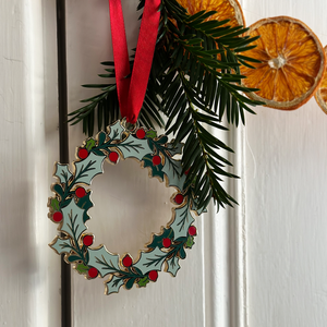 An heirloom Christmas wreath decoration with holly leaves and red berries hangs from a red ribbon on a white door handle with festive greenery and orange slice garland.