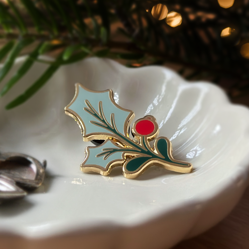 A green holly leaf pin badge with a red berry sits on a white dish with blurred greenery and lights in the background.