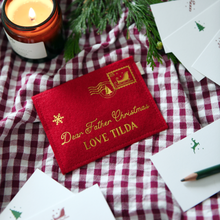 Red felt personalised Christmas envelope reading "Dear Father Christmas, Love Tilda," lying on a red gingham cloth with festive notecards, greenery, and a lit candle.