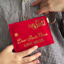 A child wearing a gingham shirt is holding a handmade red felt Christmas envelope that reads "Dear Santa Claus, Love Miles" in gold writing with a golden sleigh stamp in the top right hand corner.