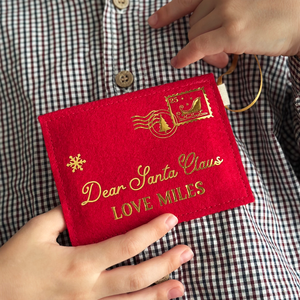 A child wearing a gingham shirt is holding a handmade red felt Christmas envelope that reads "Dear Santa Claus, Love Miles" in gold writing with a golden sleigh stamp in the top right hand corner.