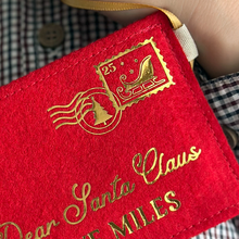 A child wearing a gingham shirt is holding a handmade red felt Christmas envelope that reads "Dear Santa Claus, Love Miles" in gold writing with a golden sleigh stamp in the top right hand corner.