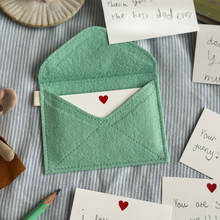 A handmade envelope lays open on light blue striped fabric. Tucked inside are mini heart notecards, with several written cards beside it. Each written card has a note to Daddy written by children.