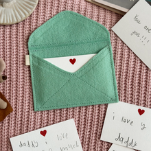 A handmade envelope lays open on pink fabric. Tucked inside are mini heart notecards, with several written cards beside it. Each written card has a note to Daddy written by children.