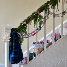 A festive Christmas garland draped over a white wooden staircase railing, featuring green pine branches, patterned paper chain links, pink ribbon bows, and a matching plaid fabric accent hanging from the banister.