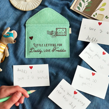 A handmade green felt envelope with the personalisation 'Little Letters For Daddy, Love Freddie' and a camping design stamp with postmark, lays on dark blue fabric. Notes to Daddy are written on the included heart notecards.