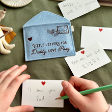 A handmade soft blue felt envelope with brown wording that reads 'Little Letters For Daddy, Love Posy' lays on a dark green fabric surface. A young child is writing a notecard to Daddy beside the envelope. Several other mini notecards are around the envelope too.