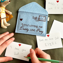 A handmade soft blue felt envelope with brown wording that reads 'Little Letters For Daddy, Love Posy' lays on a dark green fabric surface. A young child is writing a notecard to Daddy beside the envelope. Several other mini notecards are around the envelope too.