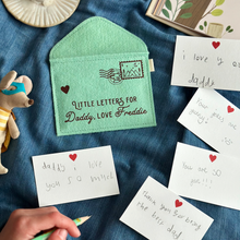 A handmade green felt envelope with the personalisation 'Little Letters For Daddy, Love Freddie' and a paper aeroplane stamp with postmark, lays on dark blue fabric. Notes to Daddy are written on the included heart notecards.