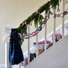 A decorated indoor staircase with a green garland draped over the banister, a paper chain garland made of patterned paper loops, a plaid scarf on the newel post, wrapped gifts, and a wicker basket.
