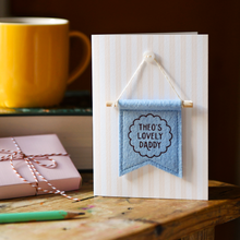 Greeting card with blue banner reading 'THEO'S LOVELY DADDY' on a wooden surface with a mug and gift box.